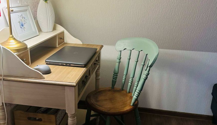 A cozy workspace featuring a vintage wooden desk with a laptop, a mouse, and a potted plant. A green wooden chair is placed at the desk. A table lamp emits a warm glow, illuminating a framed floral artwork on the wall.