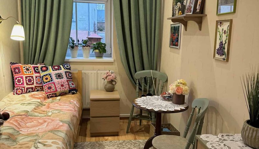 Cozy room with a single bed featuring colorful crochet pillows and a floral blanket. A small round table with chairs sits in the corner. Green curtains frame the window, and shelves with plants and photos adorn the walls. A chest of drawers is nearby.