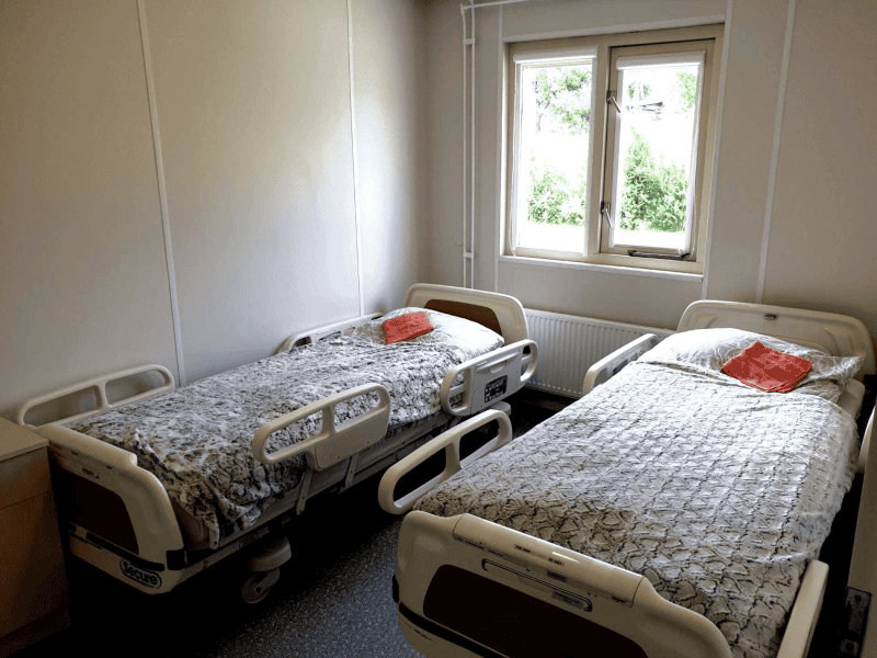 A hospital room with two empty hospital beds, each with patterned blankets and a small orange towel. The room has a window with a view of greenery outside. Walls are light-colored, and the floor is covered with speckled tiles.