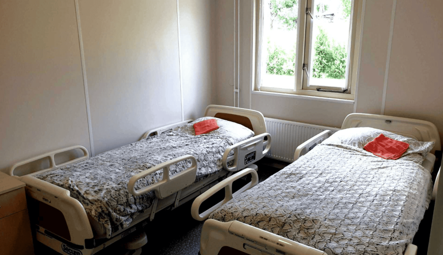 A hospital room with two empty hospital beds, each with patterned blankets and a small orange towel. The room has a window with a view of greenery outside. Walls are light-colored, and the floor is covered with speckled tiles.