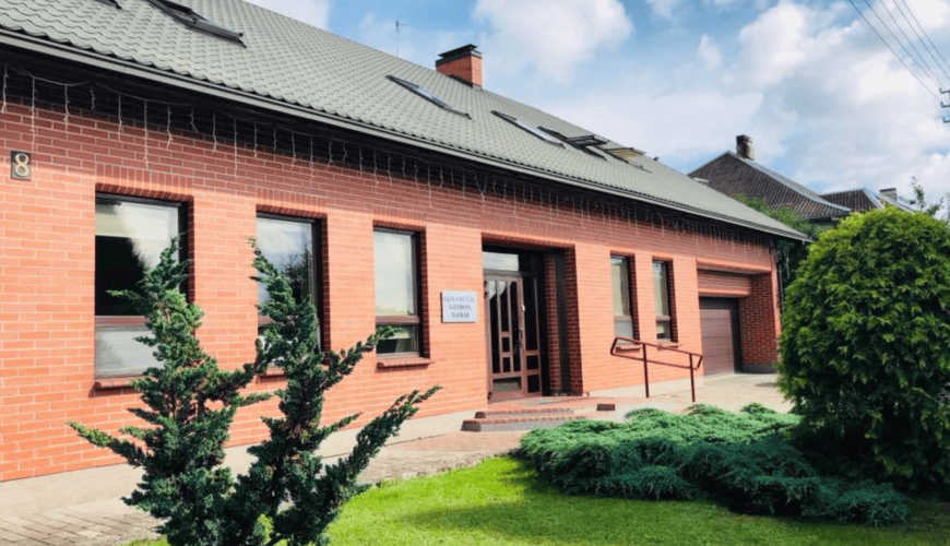A single-story red-brick building with a sloped gray roof and several windows. It has a wooden door, a small sign beside it, and a wheelchair ramp. A well-maintained lawn with shrubs is in the foreground under a partly cloudy sky.