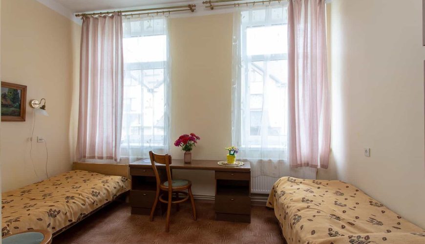 A small room with two single beds on either side, separated by a wooden desk and chair. Pink curtains cover two tall windows. A vase with red flowers and a cup with yellow flowers sit on the desk. The bedspreads have a floral pattern.
