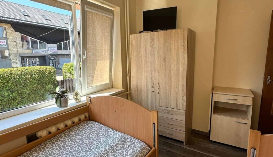 A small room featuring two wooden single beds with patterned mattresses. A window lets in natural light, illuminating a wooden wardrobe with a TV on top. A small nightstand is beside the wardrobe. The view includes a hedge and buildings outside.
