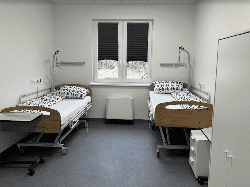 A hospital room with two empty beds featuring wood paneling, patterned bedding, overbed tables, and wall-mounted reading lamps. The room has a window with blinds, white walls, and a gray floor. A cabinet and a heater are also visible.