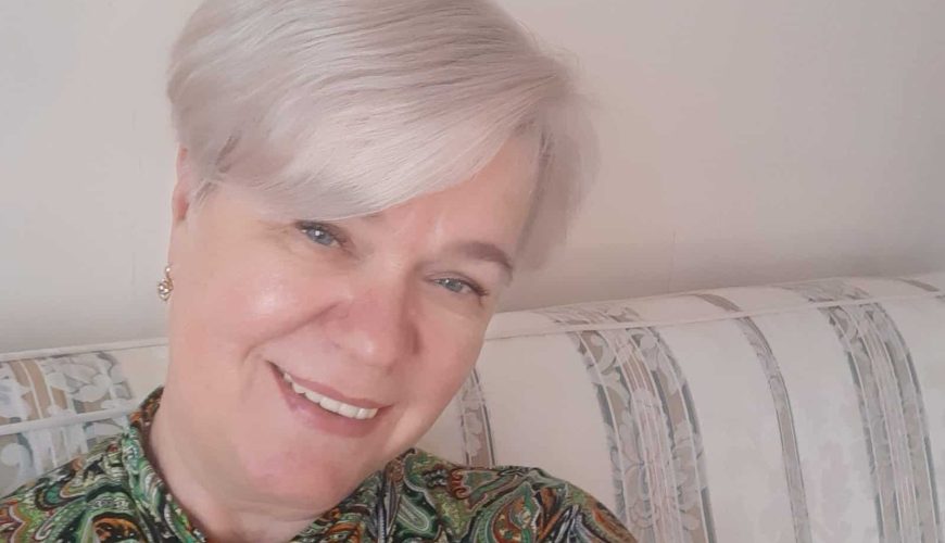 A person with short, light hair smiles at the camera. They are wearing a paisley-patterned top and are seated on a striped sofa. The background is a plain wall.
