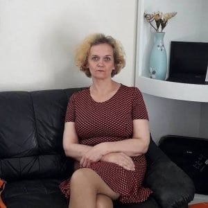 A person with short, curly hair sits on a black couch, wearing a red patterned dress. They have folded hands and a neutral expression. In the background, there's a blue vase with a decorative item and a small screen.