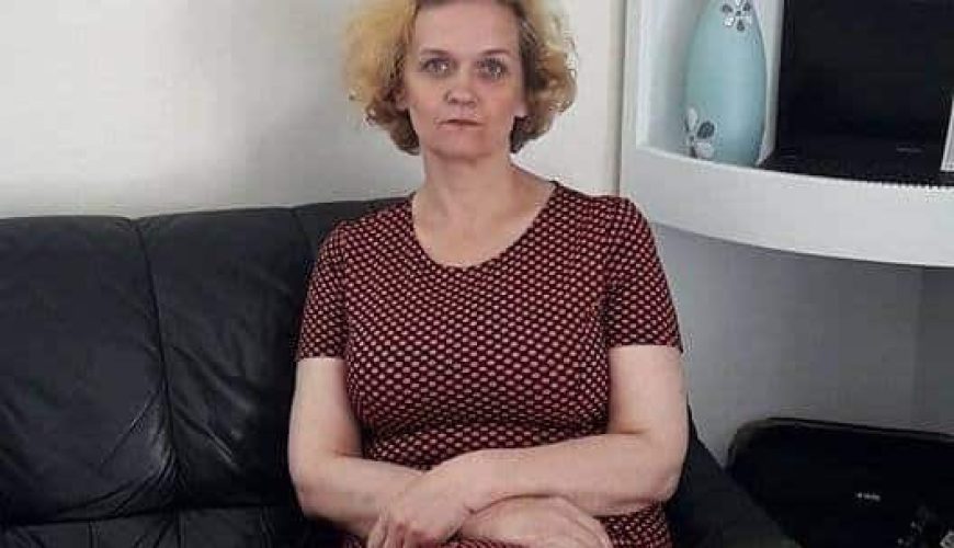A person with short, curly hair sits on a black couch, wearing a red patterned dress. They have folded hands and a neutral expression. In the background, there's a blue vase with a decorative item and a small screen.