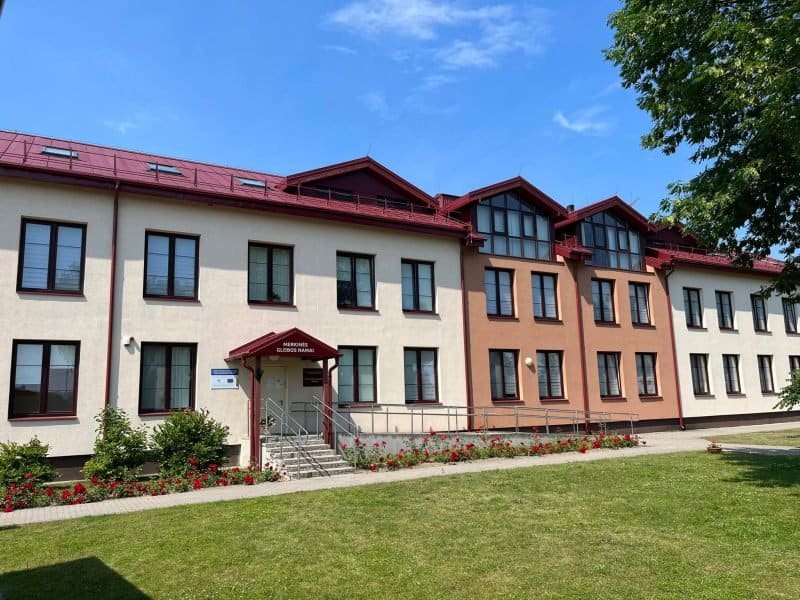 A two-story building with red roofs and cream and maroon walls stands under a blue sky. It has multiple windows and a central entrance with a ramp. The front yard features a path, a green lawn, and red flowers.