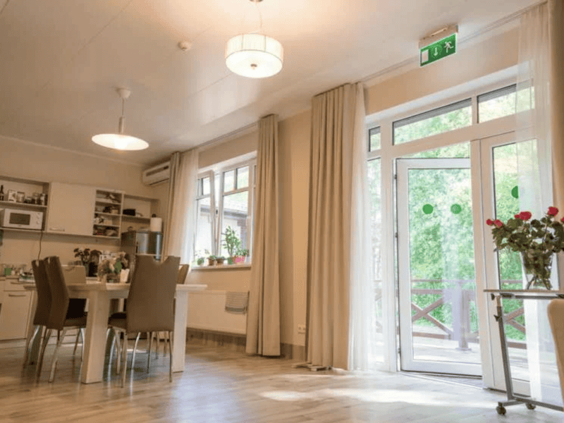 A bright, modern kitchen and dining area with a large table and chairs. The room features white cabinets, a countertop with appliances, and a vase of red flowers. Large windows and glass doors let in natural light, with views of greenery outside.