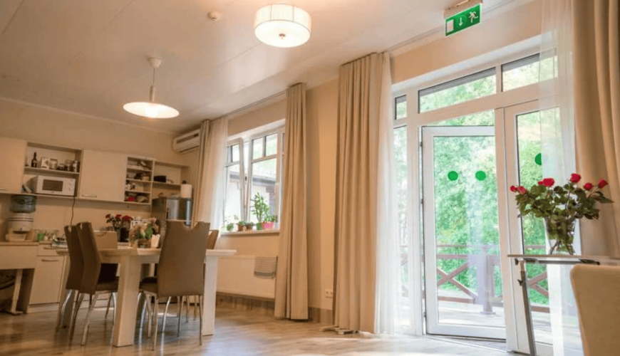 A bright, modern kitchen and dining area with a large table and chairs. The room features white cabinets, a countertop with appliances, and a vase of red flowers. Large windows and glass doors let in natural light, with views of greenery outside.