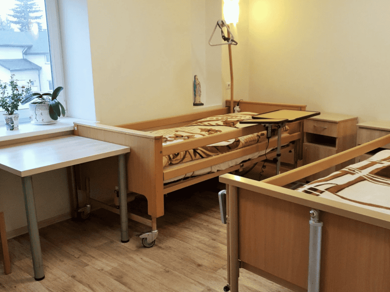 A tidy room with two adjustable hospital beds and wooden furniture. There's a window with potted plants on a table, and a lamp mounted on the wall. The flooring is laminate, and there's a serene atmosphere.