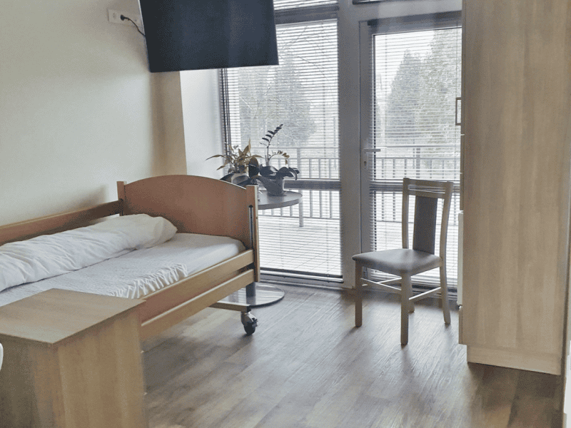 A clean, simple room with a single bed, wooden chair, small table, and a TV mounted on the wall. A large window with blinds overlooks a balcony. A plant sits on the window ledge, adding a touch of greenery to the space.