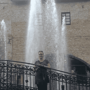 A woman stands and smiles on a decorative iron bridge in front of a tall water fountain, with a brick building and arched entrance in the background on a sunny day.