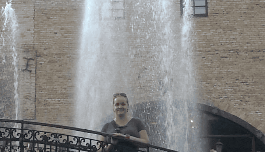 A woman stands and smiles on a decorative iron bridge in front of a tall water fountain, with a brick building and arched entrance in the background on a sunny day.