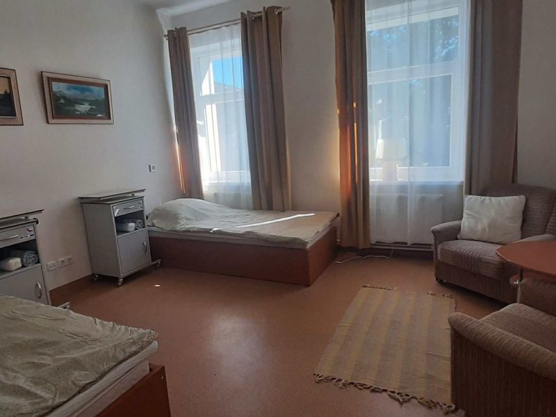 A simple room with two single beds, two nightstands, two armchairs, a small round table, a rug, and two large windows with curtains; landscape paintings hang on the white walls.