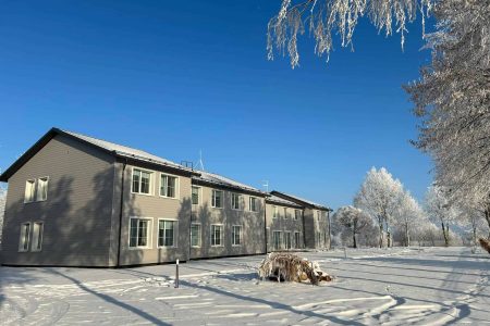 A modern two-story building stands under a clear blue sky, surrounded by snow-covered ground and trees with frosty branches on a bright winter day.
