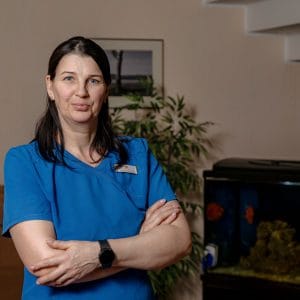 A woman in blue medical scrubs stands with arms crossed, smiling slightly. She has dark hair and wears a watch. Behind her are a green plant, a framed photo, and an aquarium against a beige wall.