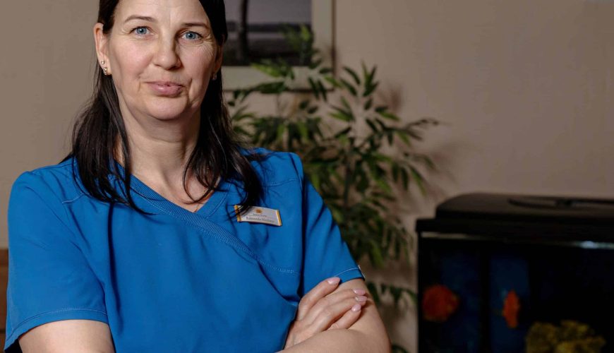 A woman in blue medical scrubs stands with arms crossed, smiling slightly. She has dark hair and wears a watch. Behind her are a green plant, a framed photo, and an aquarium against a beige wall.