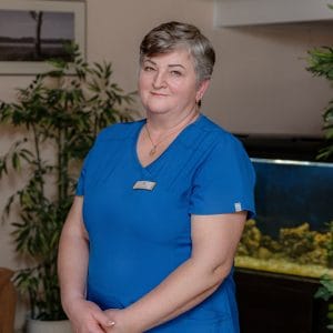 A woman with short gray hair, wearing blue scrubs and a name badge, stands indoors in front of an aquarium and potted plants, smiling gently at the camera.