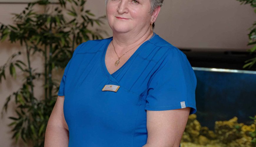 A woman with short gray hair, wearing blue scrubs and a name badge, stands indoors in front of an aquarium and potted plants, smiling gently at the camera.
