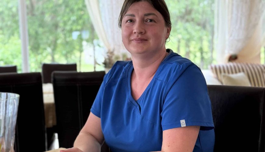 A woman wearing blue scrubs sits at a table with her hands folded, smiling softly. She is indoors, with large windows behind her revealing green trees and sheer curtains. The setting appears calm and bright.