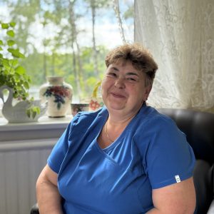 A smiling woman with short brown hair, wearing blue scrubs, sits indoors near a window with potted plants, a floral vase, and sheer curtains, with greenery visible outside.