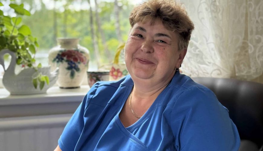 A smiling woman with short brown hair, wearing blue scrubs, sits indoors near a window with potted plants, a floral vase, and sheer curtains, with greenery visible outside.