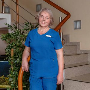 A woman with short gray hair, wearing blue medical scrubs and a name badge, stands indoors by a staircase and a potted plant, smiling at the camera.