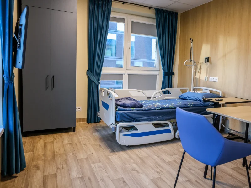 A modern hospital room with a single adjustable bed, blue bedding, a table and chair, a wardrobe, a wall-mounted TV, and large windows with blue curtains. The floor is light wood, and the walls are light-colored.