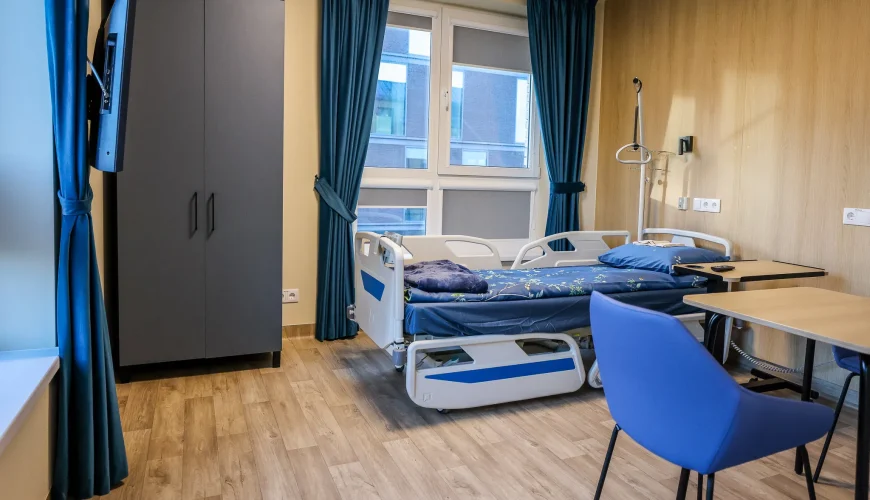 A modern hospital room with a single adjustable bed, blue bedding, a table and chair, a wardrobe, a wall-mounted TV, and large windows with blue curtains. The floor is light wood, and the walls are light-colored.