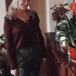 A smiling woman with long blonde hair wearing a burgundy top and plaid pants stands indoors beside a wooden sideboard decorated with potted plants and a framed photo.