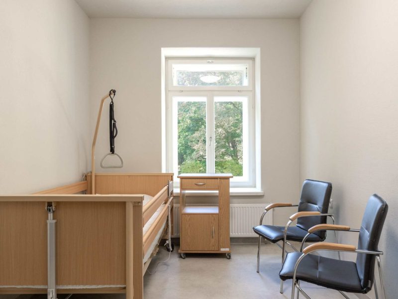 A small, bright hospital room with a wooden hospital bed, a bedside table, and two black chairs by a large window showing green trees outside. The walls are plain white, and the room appears clean and minimalistic.