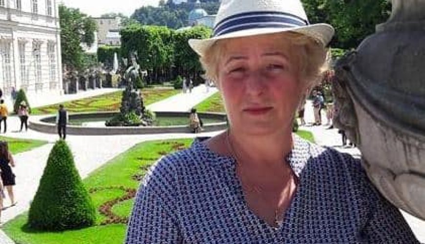 A woman wearing a white hat and patterned shirt stands next to a large stone planter in a formal garden with trimmed hedges, statues, and a distant castle on a hill in the background.