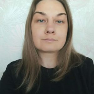 A person with straight, light brown hair and light skin is wearing a black top and looking directly at the camera, with a neutral expression. The background is light-colored with a subtle pattern.