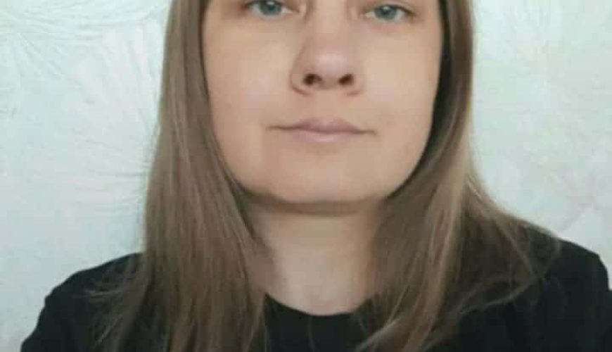 A person with straight, light brown hair and light skin is wearing a black top and looking directly at the camera, with a neutral expression. The background is light-colored with a subtle pattern.
