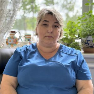 A middle-aged woman with short gray hair, wearing a blue shirt, sits indoors in front of a window with potted plants and teapots in the background, looking calmly at the camera.