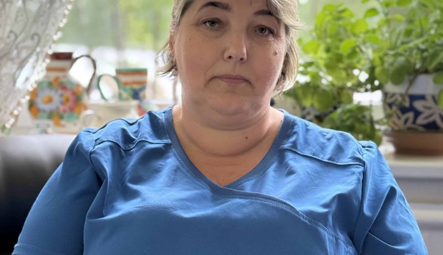 A middle-aged woman with short gray hair, wearing a blue shirt, sits indoors in front of a window with potted plants and teapots in the background, looking calmly at the camera.