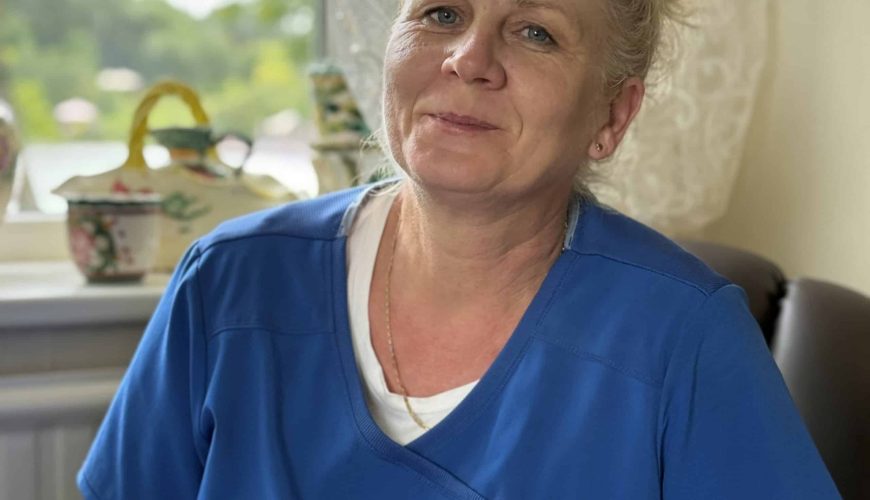 A middle-aged woman with light hair tied back, wearing blue scrubs, sits indoors near a window with lace curtains. Decorative teapots are visible on the windowsill behind her. She is smiling slightly.