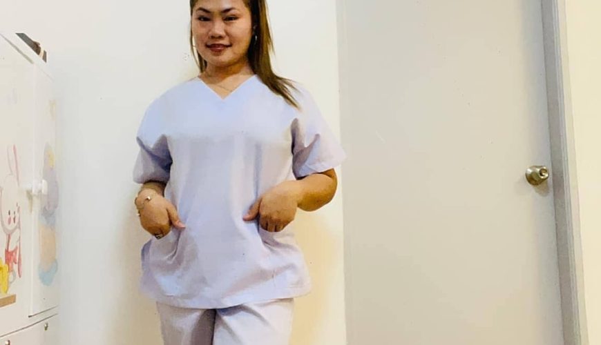 A woman wearing light purple scrubs and white sneakers stands indoors on a wooden floor, posing with her hands in her pockets in front of a white door and wall.